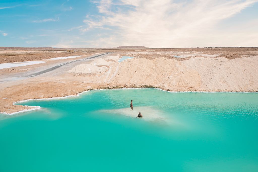 People relaxing in a surreal, crystal-blue salt lake in the Siwa Oasis surrounded by desert sands. A peaceful, non-touristy thing to do in Egypt and an Egypt bucket list essential.