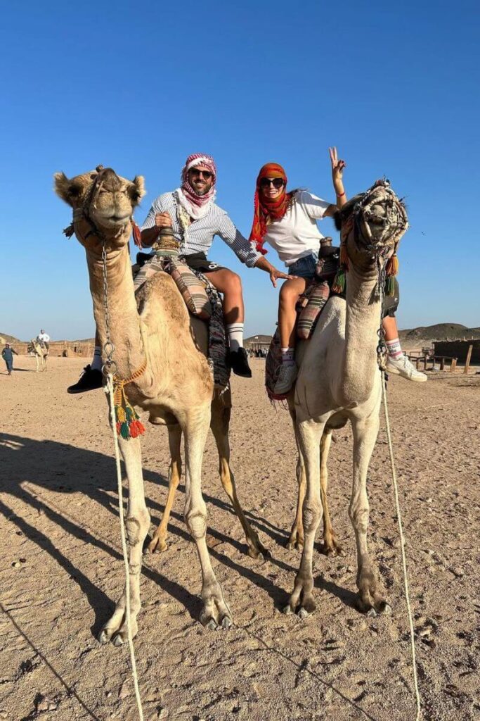A smiling couple wearing headscarves and relaxed desert attire rides camels under the clear blue sky. Their outfits reflect practical Egypt outfit ideas and align with the Egypt dress code for desert adventures, showing what to wear in Egypt for women and men.