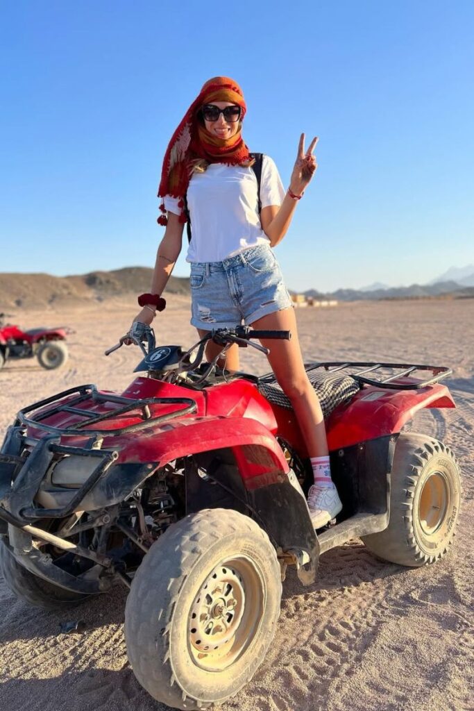 A woman poses on a red quad bike wearing a white t-shirt, denim shorts, and a red headscarf with desert mountains in the background. While casual, her look represents active Egypt outfit ideas and sparks conversation about the Egypt dress code and what to wear in Egypt for women on outdoor excursions.