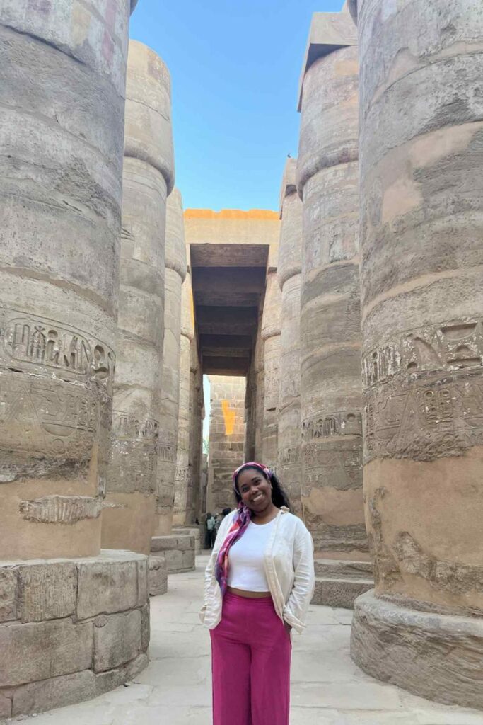 Smiling among the massive carved columns of Karnak Temple, a woman wears fuchsia pants, a white crop top, and a light jacket. This stylish and respectful look fits the Egypt dress code and gives ideas for what to wear in Egypt women love.