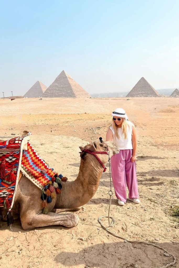 A woman dressed in loose pink pants, a white tank top, and a headscarf stands beside a camel with the pyramids of Giza in the background. Her outfit is a perfect example for any Egypt packing list and shows how to dress in Egypt respectfully and comfortably.