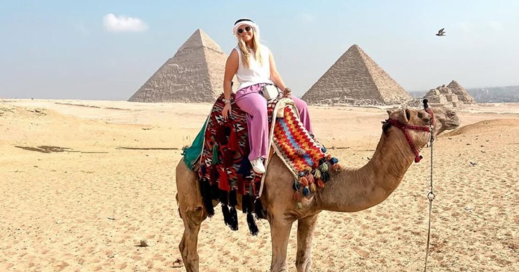Smiling atop a colorfully decorated camel, a woman wears modest and breathable travel clothes with the pyramids of Giza in the background. This outfit is a great example from an Egypt packing list women should follow, especially when considering what to pack for Egypt and how to stay cool while packing for Egypt desert adventures.