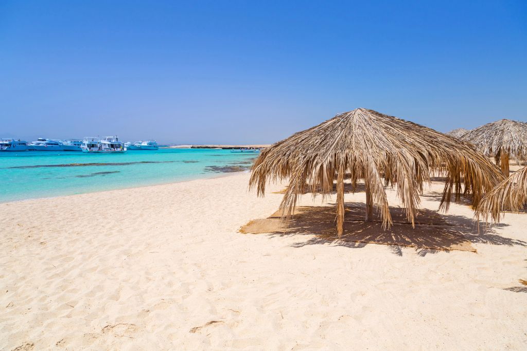 Thatched umbrellas cast shadows on a quiet white sand beach with turquoise water and boats in the background in Hurghada. A beach escape often added to a 10 day Egypt itinerary or Egypt travel guide for coastal relaxation.