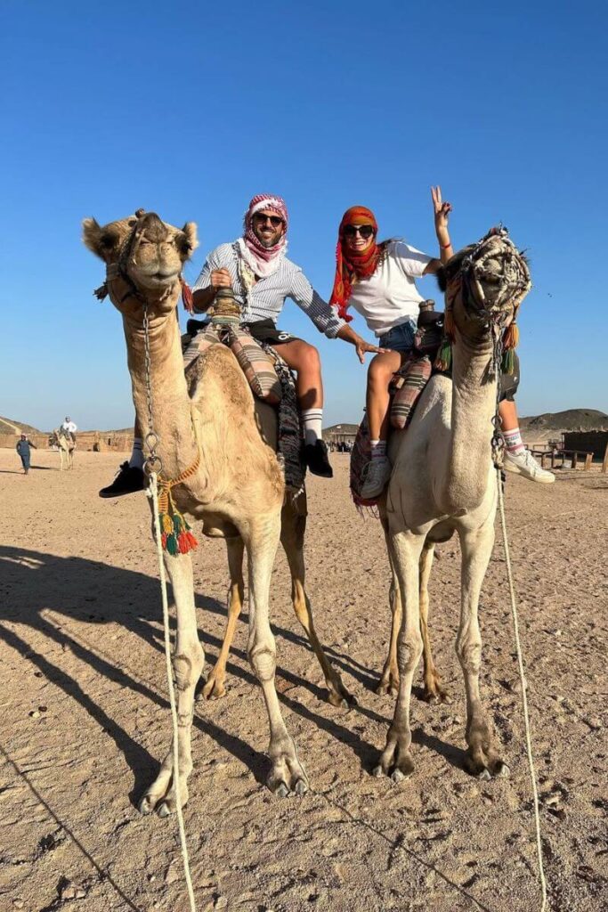 Two travelers riding camels through the golden desert under clear blue skies, flashing peace signs. Camel rides are a classic must-do in Egypt and one of the most unique things to do in Egypt.