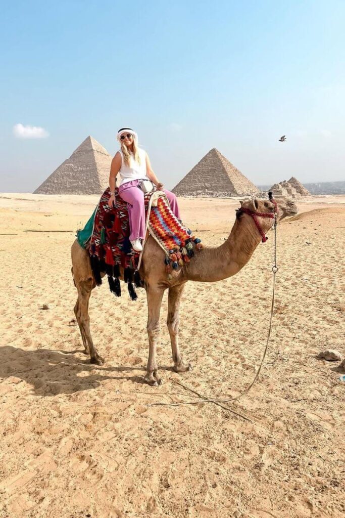 Traveler posing on a decorated camel with the iconic pyramids of Giza rising behind her under a clear blue sky. Camel rides by the pyramids are a classic Egypt bucket list experience and a must-do in Egypt for first-time visitors.