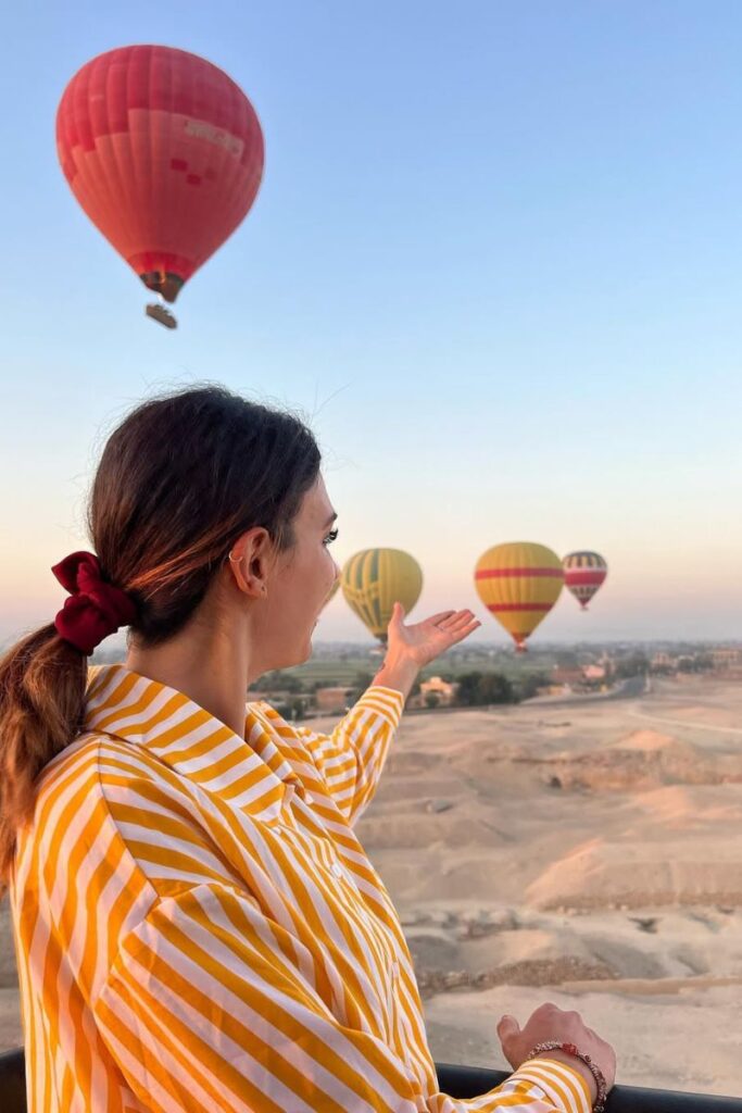 A woman in a yellow and white striped shirt smiles and gestures toward colorful hot air balloons rising at sunrise. Her breezy outfit fits the Egypt dress code and is a stylish example of what to wear in Egypt for women looking for comfortable yet cute Egypt outfit ideas.