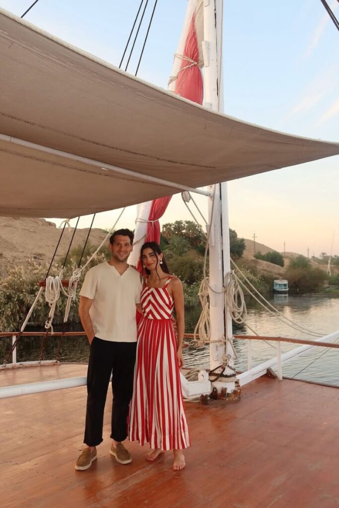 A couple poses on the wooden deck of a sailboat on the Nile, the woman in a red-striped dress and the man in casual travelwear. These relaxed yet modest outfits show what to pack for Egypt in November and suit any Egypt packing list for couples.