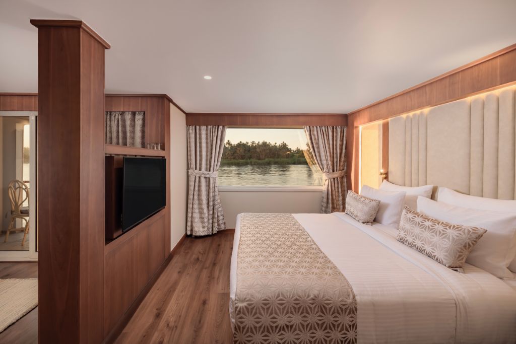 A spacious Nile cruise cabin with large windows, a king-size bed, and neutral decor. Add sleepwear and evening resort attire to your Egypt travel packing list for moments like these when cruising the Nile in comfort.