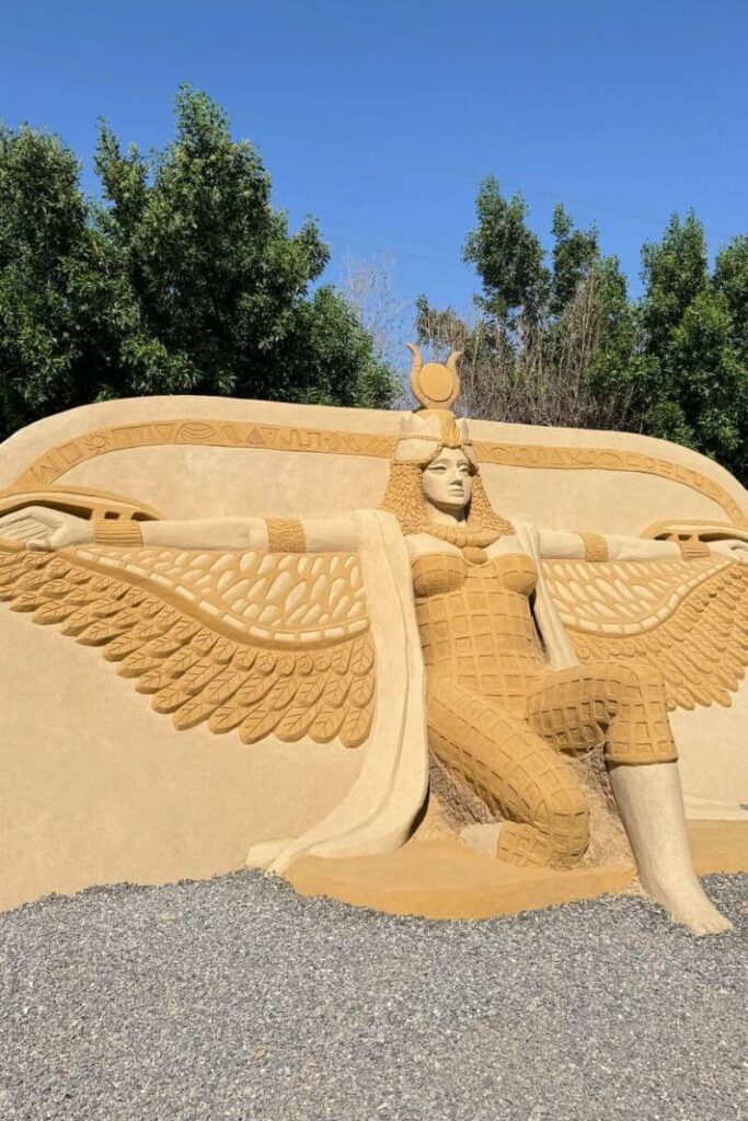 Large sand sculpture of an ancient Egyptian goddess with outstretched wings, carved in detail against a backdrop of trees and blue sky. This artistic display is one of the unique things to do in Hurghada Egypt and makes a great stop for culture lovers browsing a Hurghada travel guide.