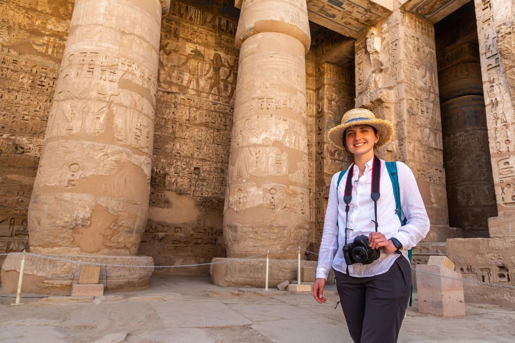 A woman wearing a straw hat, long-sleeve shirt, and cargo pants smiles in front of massive hieroglyph-covered pillars at an ancient Egyptian temple. Her outfit is a solid choice for any Egypt packing list and shows how to dress in Egypt for temple visits.