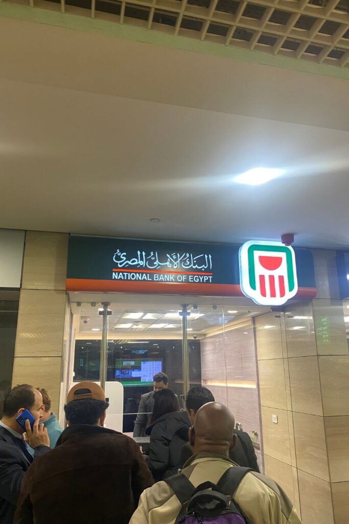 Tourists exchanging money at the National Bank of Egypt inside an airport – a key moment for those seeking tips for visiting Egypt, understanding the cost to travel to Egypt, and advice for travelers on exchanging money safely.