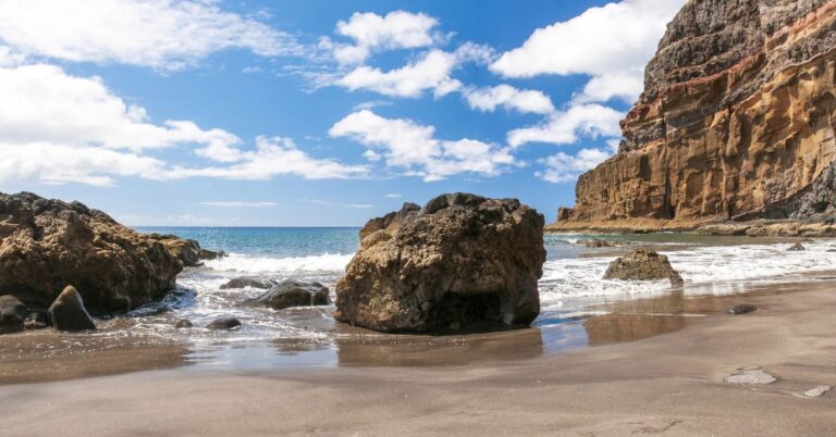 A rugged volcanic beach with dark sand, scattered rocks, and clear waves lapping the shore under a bright blue sky. This dramatic coastline is part of the Canary Islands, one of the best warm destinations in October for beach lovers and a top choice for travelers asking where is warm in October in Europe.