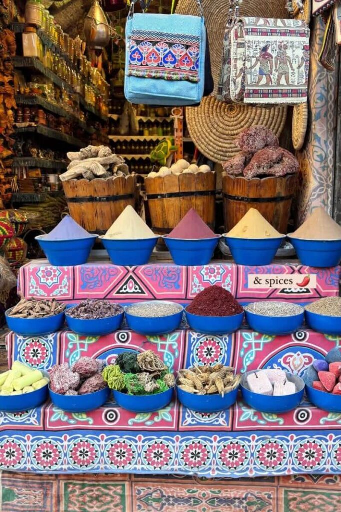 Colorful bowls of spices and dried goods line a vibrant Egyptian market stall, with souvenir bags hanging above. Add a small tote bag and cash to your packing list for Egypt woman, and don’t forget essentials for browsing markets on your Egypt travel packing list.