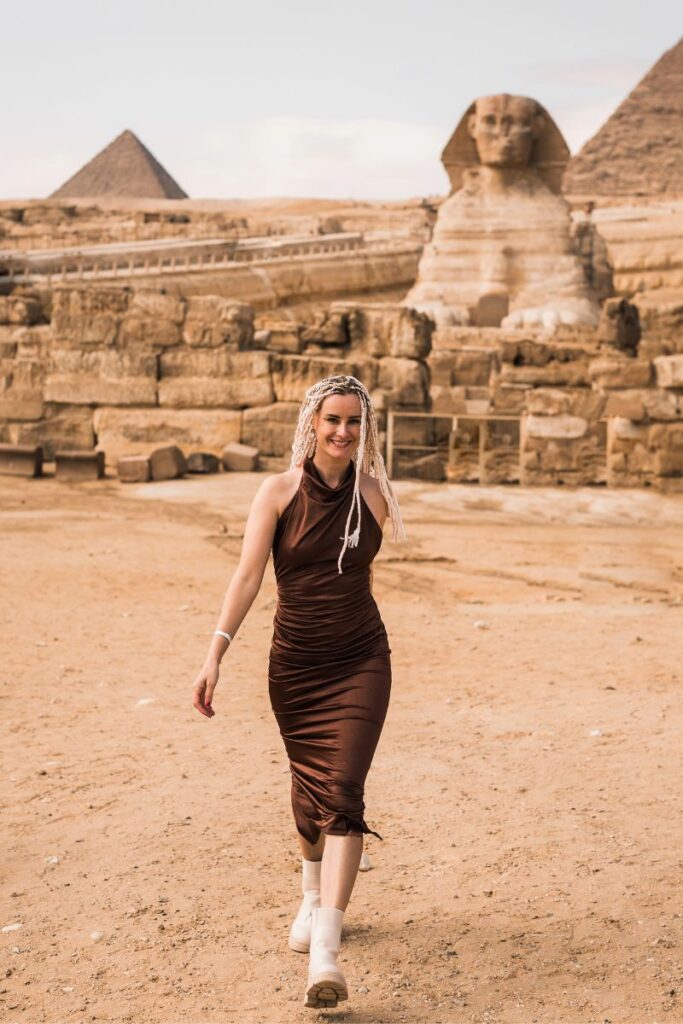 Smiling woman in a brown dress walking near the ancient ruins of the Sphinx with pyramids in the background. Exploring this area is a classic Egypt experience and one of the most iconic things to do in Egypt.