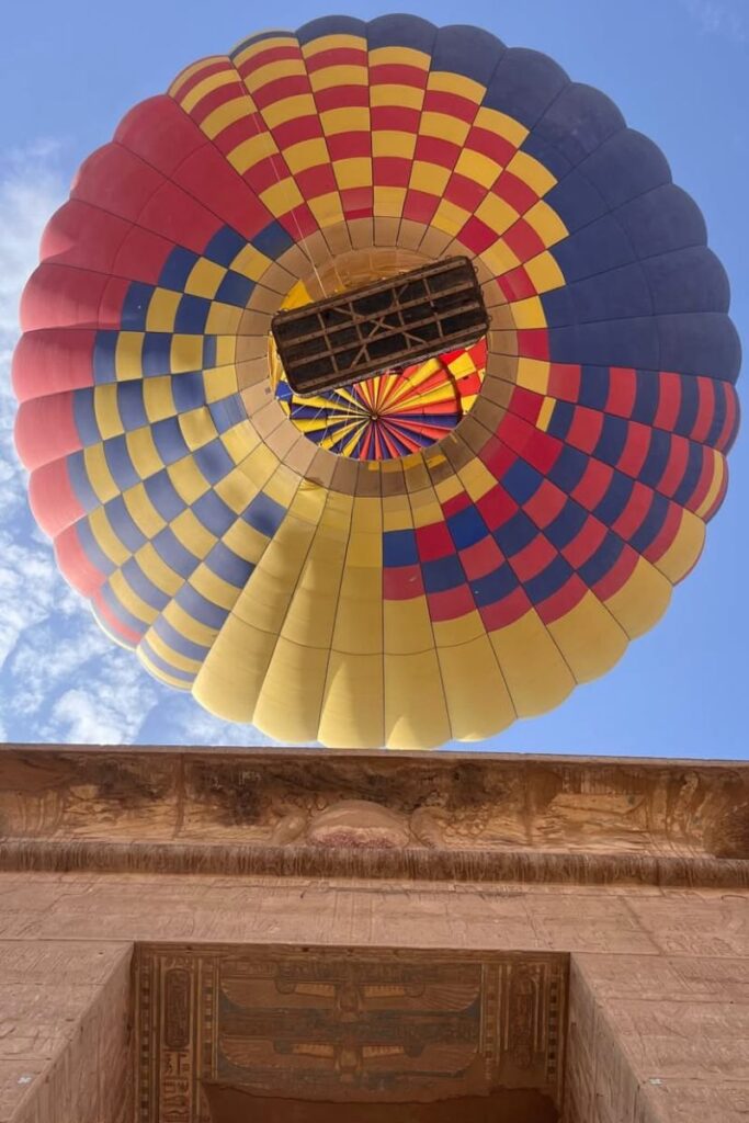A hot air balloon with red, yellow, and blue panels floats directly overhead above temple ruins in Egypt. Comfortable layers for sunrise excursions and secure shoes belong on every packing list for Egypt and are key for adventurous activities in your Egypt travel packing list.