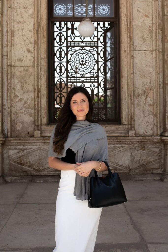 A woman dressed modestly in a white skirt, black top, and gray shawl stands near a historic mosque with intricate window designs. Her outfit is a great example of how to dress in Egypt and the appropriate dress code in Egypt for women.