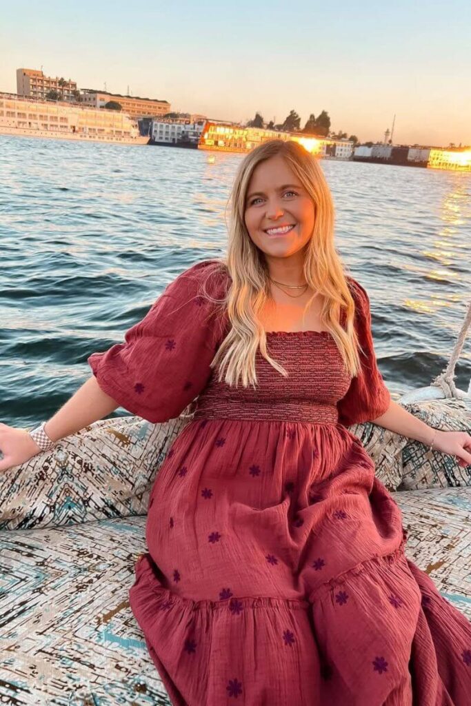 Seated on a patterned sailboat at sunset, a woman in a flowing maroon dress enjoys the Nile River. Her outfit highlights what to wear in Egypt in November and aligns with Egypt clothes that balance comfort and culture.
