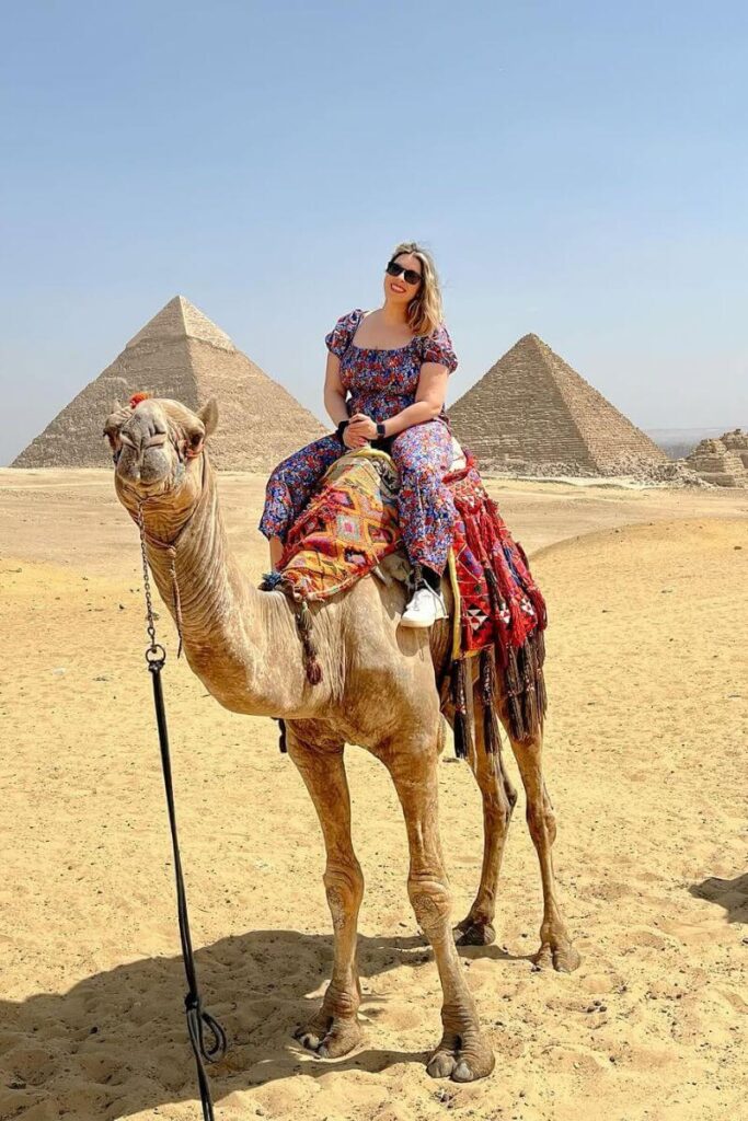 A woman sits atop a decorated camel in front of the pyramids wearing a floral jumpsuit and sunglasses. This is a practical yet fun example of what to wear to the pyramids in Egypt and follows the Egypt dress code for tourists.