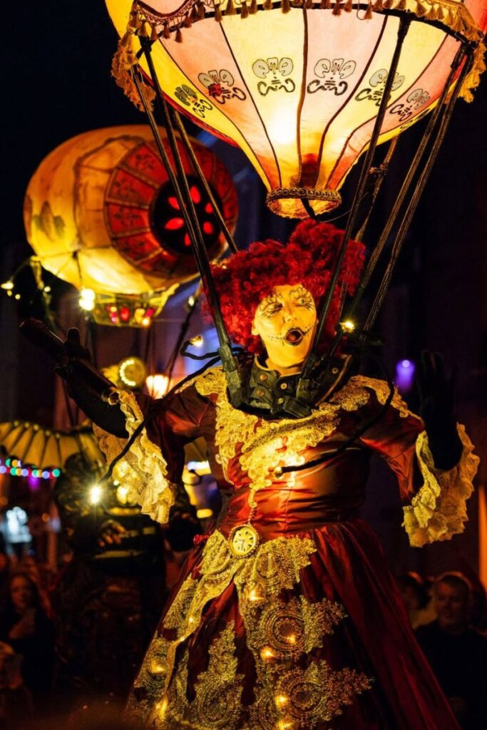 A costumed performer in a red Victorian gown with gold embroidery and glowing lights wears a large illuminated balloon headpiece, entertaining a crowd at night during a Halloween event, perfect for fun Halloween vacation ideas.
