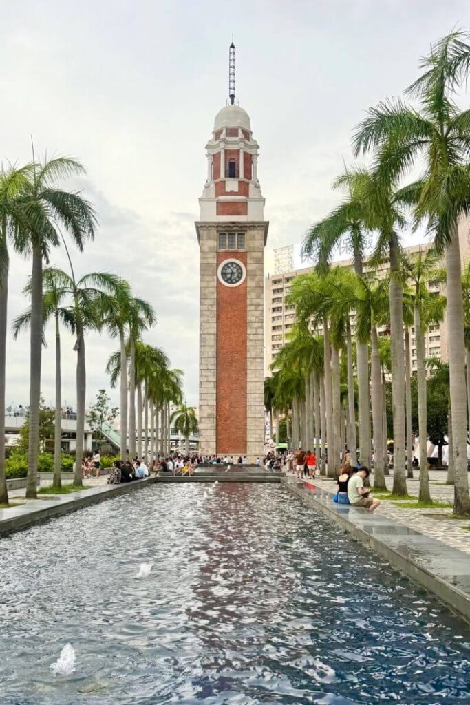 A tall brick and stone Clock Tower in Tsim Sha Tsui, one of the most recognizable Hong Kong landmarks, stands at the end of a palm-lined reflecting pool with people relaxing along the water’s edge. This scenic spot is one of the top things to do in Hong Kong and a great stop for anyone planning a 1 day in Hong Kong itinerary.