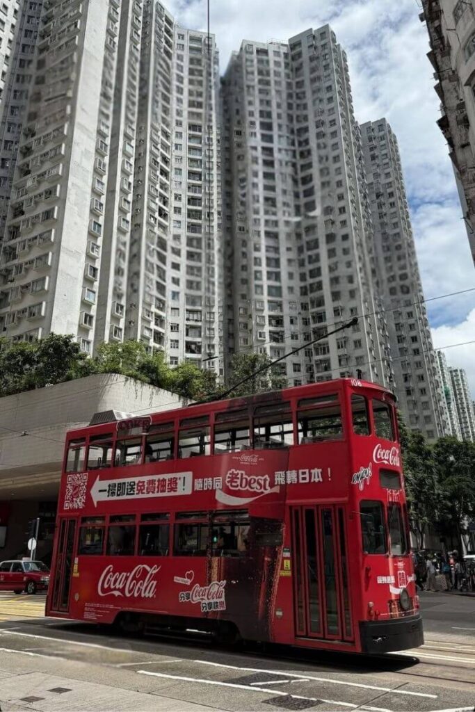 A Coca-Cola-branded red double-decker tram rolls past towering residential buildings on a sunny day. This classic tram ride is one of the iconic things to do in Hong Kong island and fits well into any Hong Kong landmarks list.