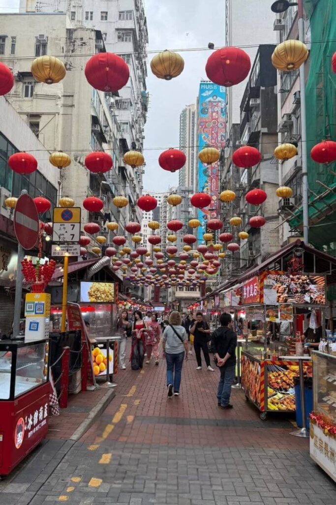 A bustling street market with rows of red and gold lanterns overhead and colorful food stalls on both sides, drawing locals and tourists alike. Visiting markets like this is one of the most authentic things to do in Hong Kong China and a favorite among unusual things to do in Hong Kong.