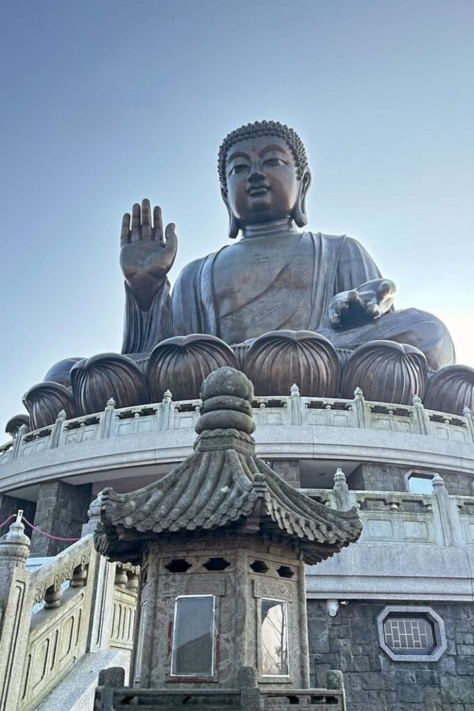 A low-angle view of the Tian Tan Buddha statue with one hand raised in blessing, sitting atop a circular podium with stone carvings. This peaceful monument on Lantau Island is one of the top things to do in Hong Kong and makes for a serene day trip from Hong Kong.