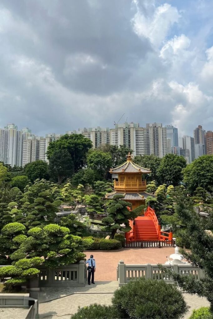 A peaceful scene of the golden Pavilion of Absolute Perfection and its red bridge nestled among manicured trees in Nan Lian Garden, with high-rise buildings in the background. This tranquil park is one of the most unique places to visit in Hong Kong and perfect for those seeking cool things to do in Hong Kong away from the crowds.