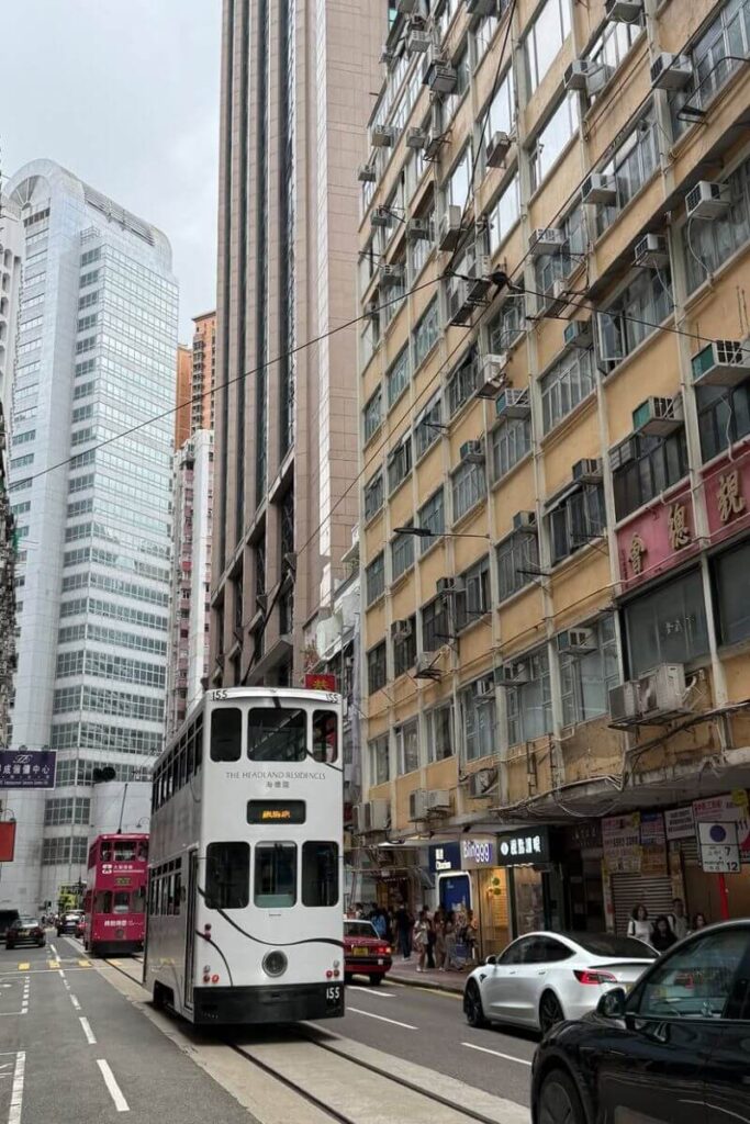 A white double-decker tram labeled "The Headland Residences" moves through a narrow street flanked by aging buildings and modern high-rises. Riding the tram is among the top things to do in Hong Kong Island and a fun way to explore the city on a 1 day in Hong Kong itinerary.