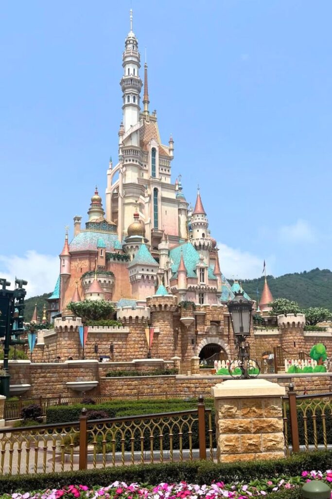 The colorful castle at Hong Kong Disneyland with turquoise rooftops and mountain views behind. A magical attraction for families debating Hong Kong or Singapore vacations.