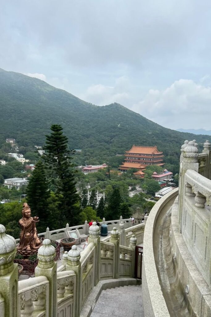 A scenic mountain view from the base of the Tian Tan Buddha shows visitors, a bronze offering statue, and the red-roofed Po Lin Monastery in the distance. This peaceful spot is one of the best places to visit in Hong Kong and perfect for day trips from Hong Kong.