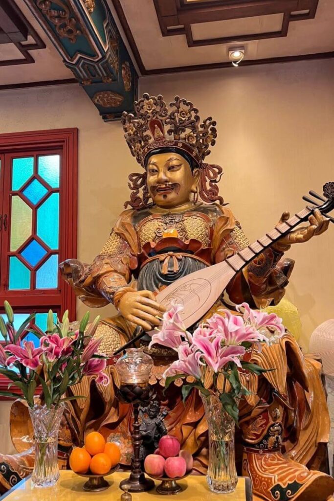 An ornate statue of a deity holding a lute is surrounded by offerings of flowers, oranges, and peaches inside the richly decorated Wong Tai Sin Temple. This temple is one of the most spiritual and cultural things to do in Hong Kong and among the top Hong Kong landmarks.