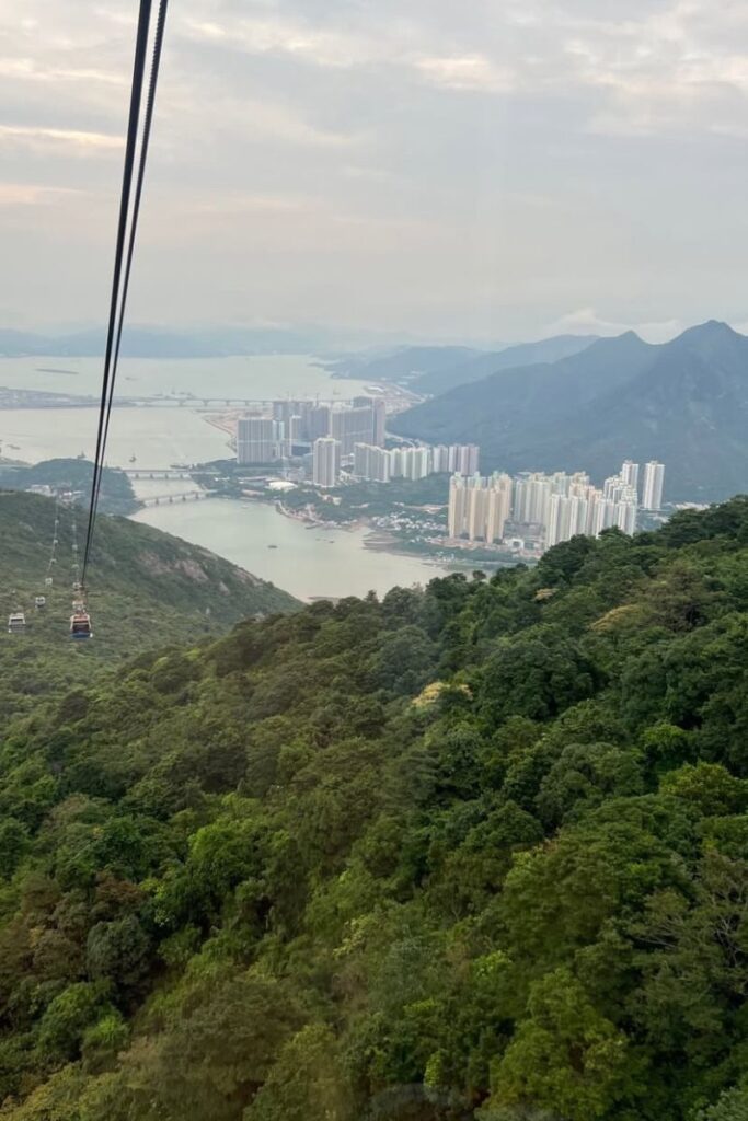 Aerial view from inside the Ngong Ping 360 cable car, soaring above lush green mountains with views of the coastline and high-rise buildings in the distance. This scenic ride is one of the top day trips from Hong Kong and one of the most fun things to do in Hong Kong for panoramic views.