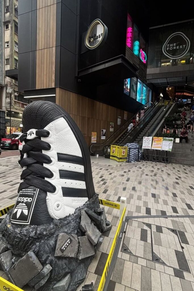 A giant black-and-white Adidas sneaker sculpture sits outside The Forest shopping center in Mong Kok, surrounded by city signage and escalators. Visiting this area is one of the most cool things to do in Hong Kong and a hotspot for streetwear shopping in Hong Kong.