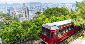 A red Peak Tram ascends through lush greenery with a panoramic view of Victoria Harbour and Hong Kong Island’s skyline in the background. Riding the Peak Tram is one of the best things to do in Hong Kong and a must-have experience on any Hong Kong itinerary.