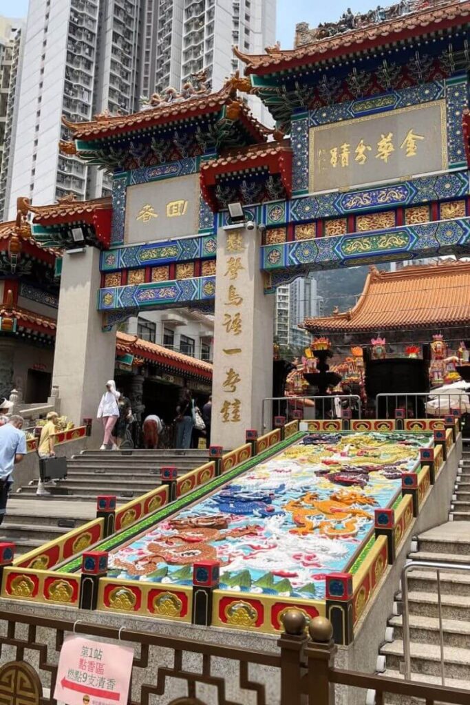 The entrance of Wong Tai Sin Temple features richly detailed Chinese architecture, gold accents, and a colorful dragon mural at the steps, with high-rise buildings in the background. A spiritual and cultural hub, this temple is one of the most meaningful places to visit in Hong Kong and a highlight among Hong Kong attractions.