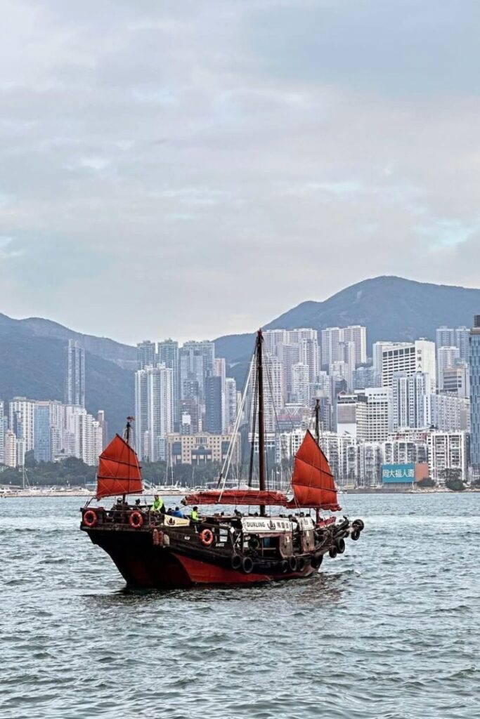 A traditional junk boat with red sails glides across Victoria Harbour, surrounded by modern skyscrapers and mountains. Staying in hotels in Hong Kong with harbour view offers travelers a chance to enjoy this classic scene, making it one of the best areas to stay in Hong Kong for tourists and families.
