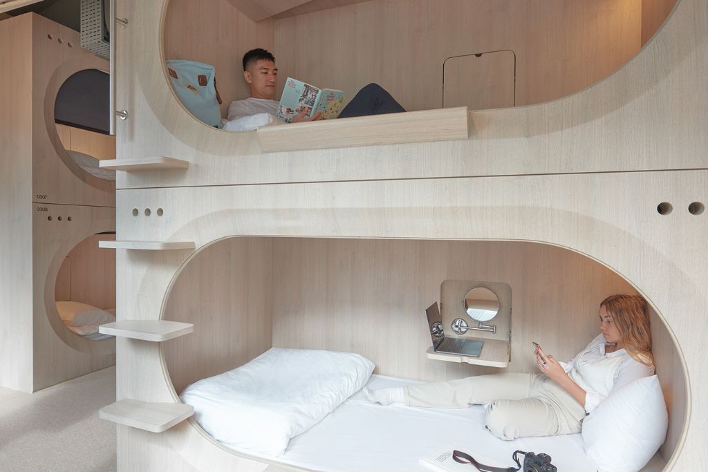 Modern capsule hotel in Singapore with cozy wooden pods, where a man reads a comic book in the upper bunk and a woman lounges with her phone in the lower pod. These stylish capsule accommodations are among the most affordable and trendy places to stay in Singapore for solo travelers and digital nomads.