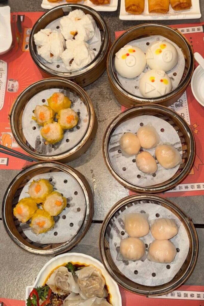 An overhead view of bamboo baskets filled with dumplings, siu mai, and bao buns at a bustling restaurant. A delicious highlight when deciding between Hong Kong or Singapore food experiences.