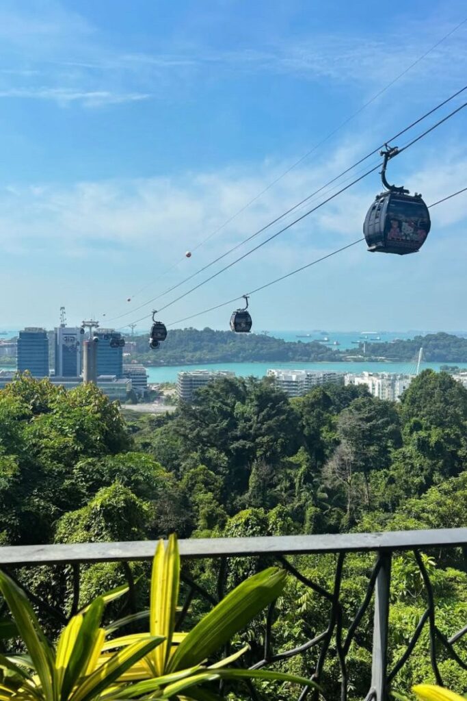 Cable cars glide over lush greenery toward Sentosa Island, with the sparkling blue sea and skyline in the distance. Riding the cable car is one of the most scenic activities in Singapore and a popular way to explore attractions near the coast.