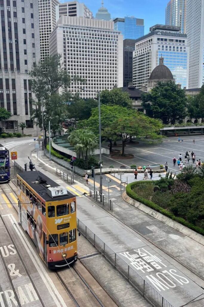 A yellow double-decker tram travels through Hong Kong’s Central district surrounded by skyscrapers and city parks. Riding the tram is a classic tourist tip for Hong Kong and an affordable way to explore the city.