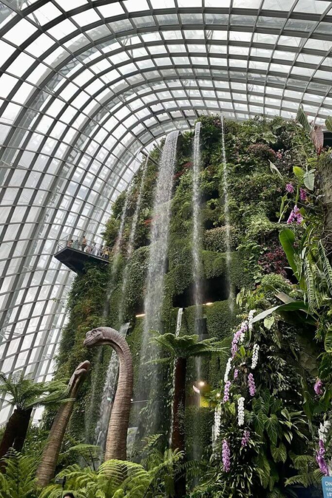 A massive indoor waterfall cascades down a green-covered mountain inside the Cloud Forest dome at Gardens by the Bay, surrounded by tropical plants and dinosaur sculptures. This indoor attraction is one of the best indoor things to do in Singapore and a must-see for nature lovers.