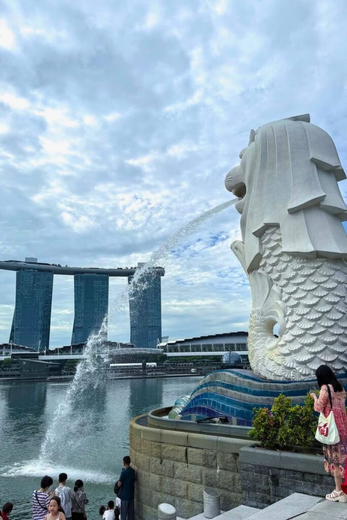 The famous Merlion statue sprays water into Marina Bay with Marina Bay Sands in the distance and tourists gathered around. Visiting this iconic site is one of the most essential Singapore things to do and a top photo stop for visitors.