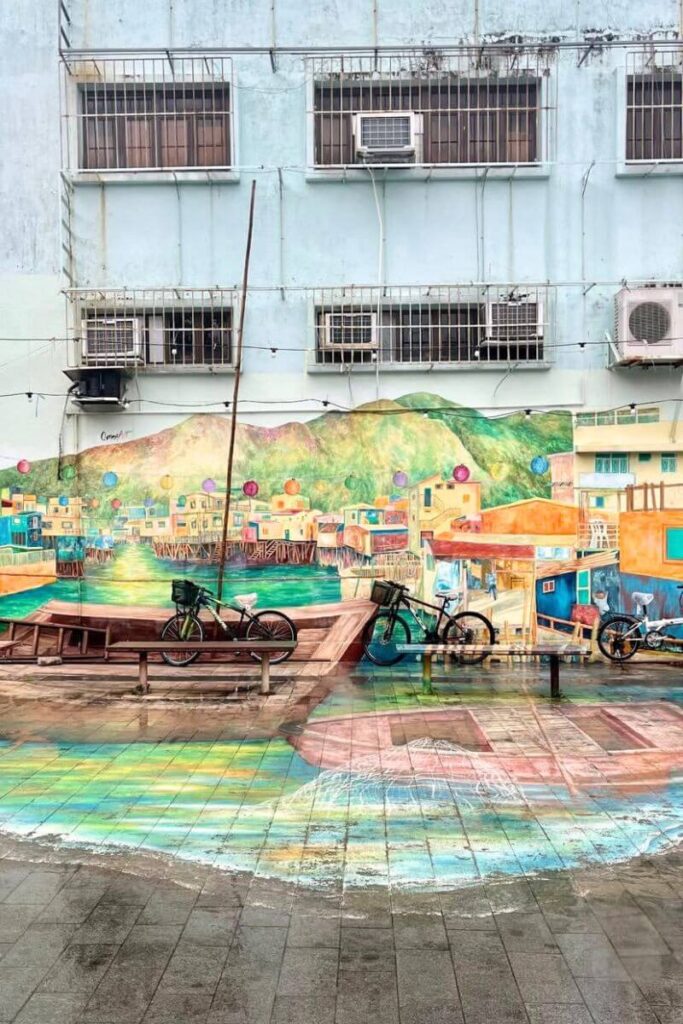 A vibrant street mural in Tai O fishing village features painted stilt houses, water reflections, and two real bicycles parked in front. Visiting Tai O is one of the most unique things to do in Hong Kong and makes for a memorable day trip from Hong Kong.