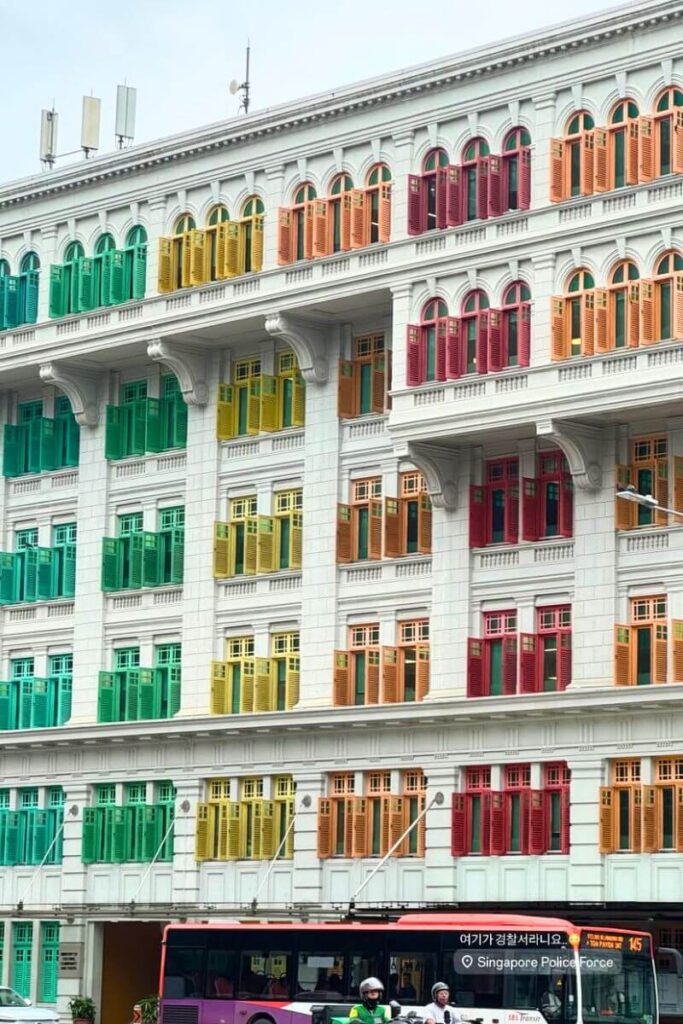 A white colonial-style building with rainbow-colored window shutters, known as the Old Hill Street Police Station, stands above passing buses and motorbikes. This eye-catching architectural landmark is one of the most unique things to do in Singapore for architecture and photography fans.