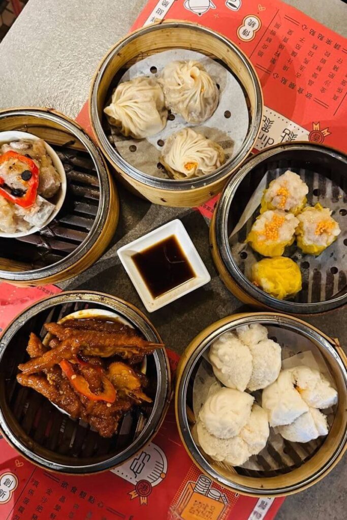 Top-down view of classic dim sum dishes including siu mai, xiao long bao, chicken feet, and steamed buns served in bamboo steamers with dipping sauce. Sampling dim sum is one of the top activities in Hong Kong and a can't-miss for any 1 day in Hong Kong food adventure.