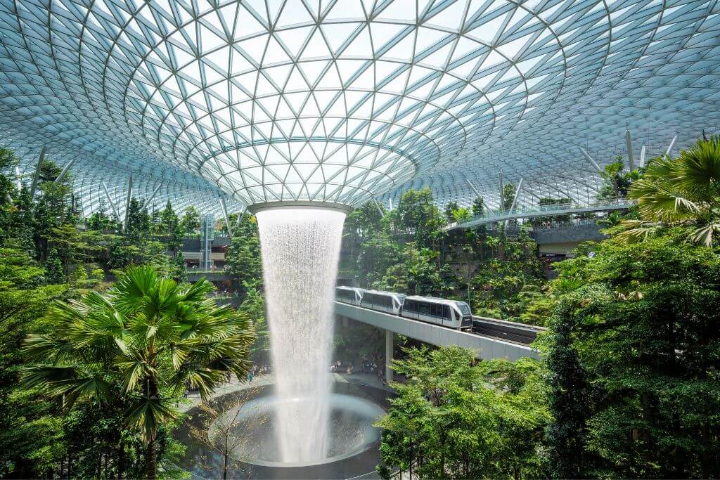 The Rain Vortex waterfall cascades from the glass-domed ceiling of Jewel Changi Airport, surrounded by lush indoor greenery and a passing skytrain. This world-renowned attraction ranks among the top things to do in Singapore airport and is one of the most unique indoor things to do in Singapore.