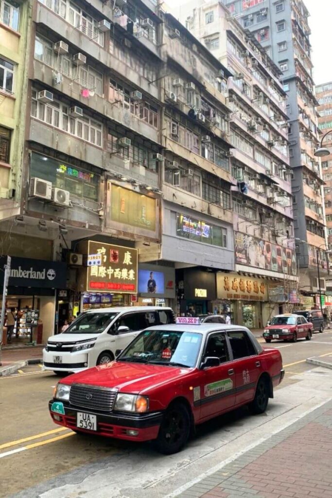 Classic red Hong Kong taxi parked along a busy street lined with old apartment buildings and neon signs. A must-know Hong Kong tourist tip is that red taxis operate mainly in Kowloon and Hong Kong Island.