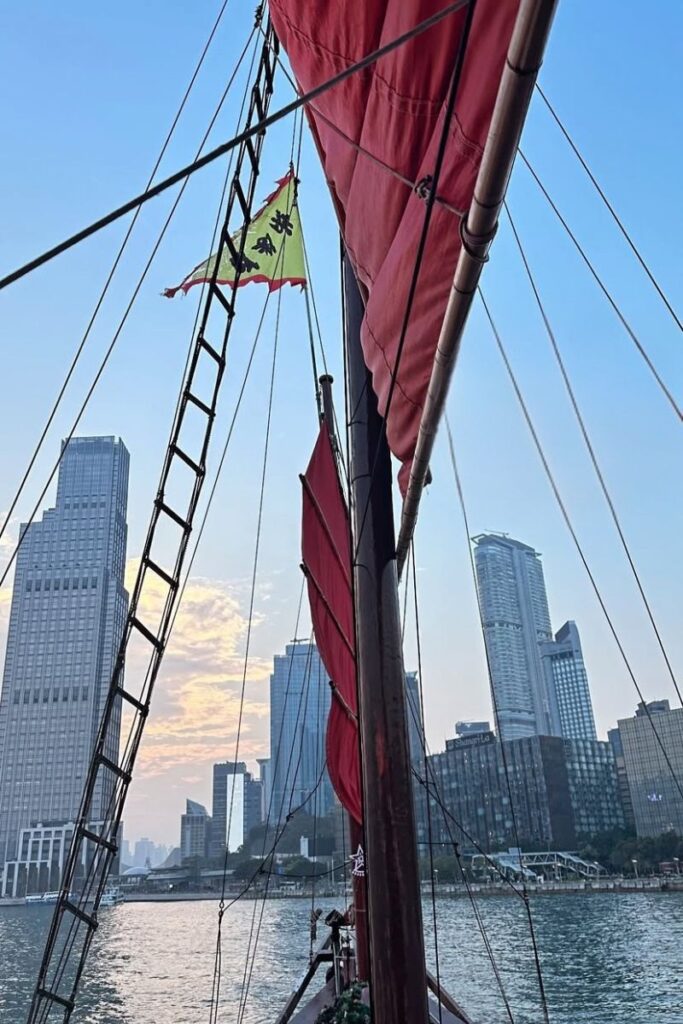 Perspective from onboard a red-sailed junk boat on Victoria Harbour, with skyscrapers and a soft sunset in the background. Sailing in a junk boat is one of the most unique things to do in Hong Kong and perfect for a scenic 1 day in Hong Kong experience.