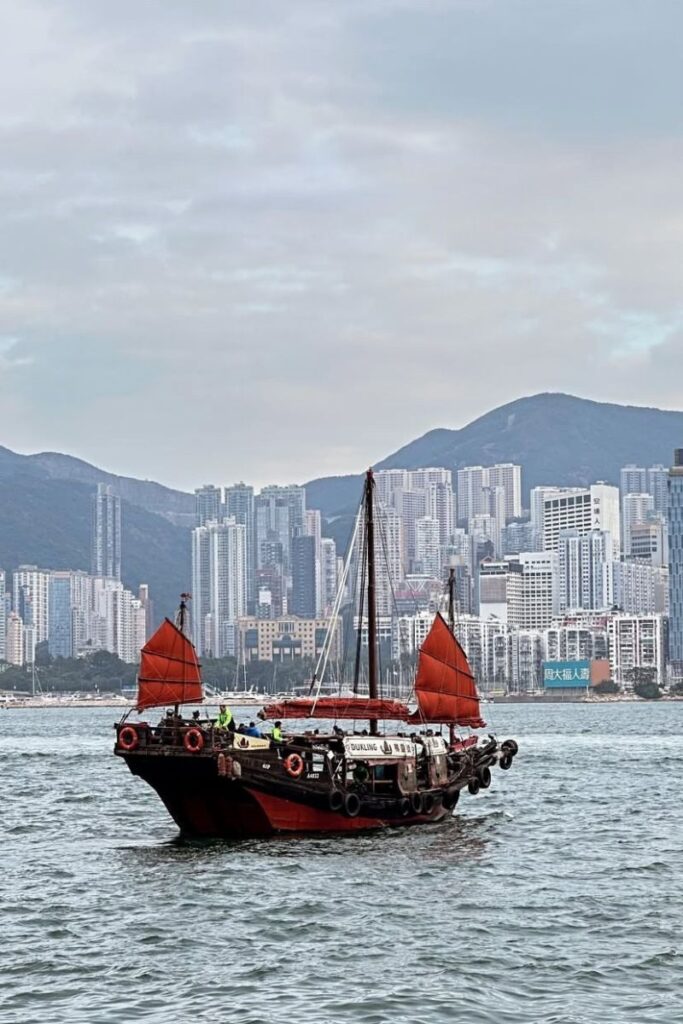 A traditional red-sailed junk boat cruises across Victoria Harbour with Hong Kong’s high-rise skyline and mountains in the background. Sailing in a junk boat is one of the most iconic things to do in Hong Kong and a scenic highlight for any 1 day in Hong Kong itinerary.